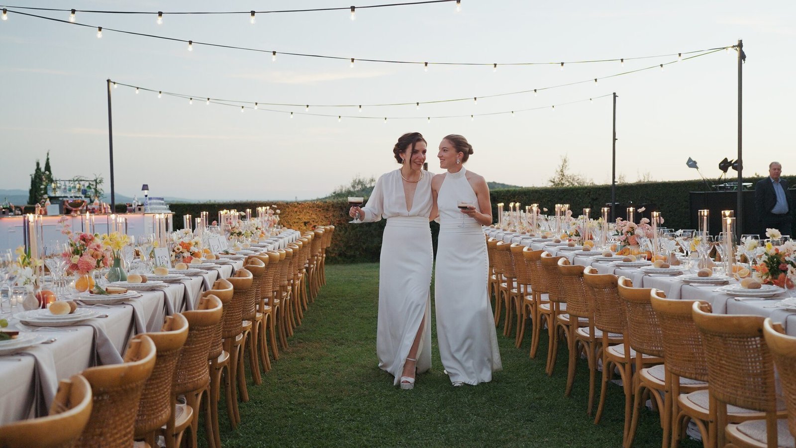 Two brides walking between long reception tables under string lights at dusk at Castello di Vicarello